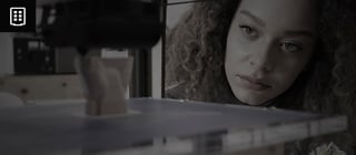 Foto de uma jovem observando atentamente uma impressora 3D em ação. O foco da atenção dela está no objeto sendo impresso